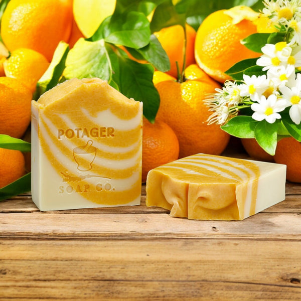 Two bars of orange dreamsicle soap with 'Potager Soap Co.' branding in front of oranges and ylang ylang flowers on a wooden surface.