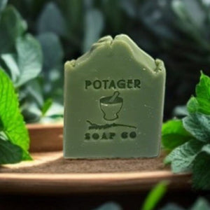 Eucalyptus Mint soap bar in green by 'Potager Soap Co' branding on a wooden stand surrounded by Eucalyptus and mint leaves.