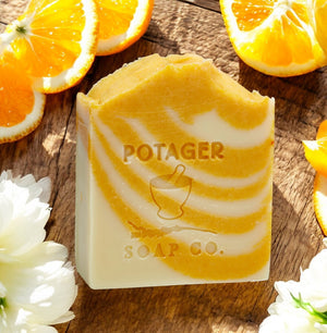 Bar of orange dreamsicle soap with 'POTAGER SOAP CO.' branding on a wooden surface with orange slices and ylang ylang flowers.