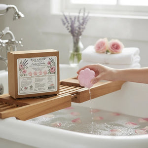 Potager Soap Co. Secret Garden flower-shaped pink bath bomb being held over a bathtub with rose petals in the water.