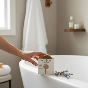Hand holding a Potager Soap Co. lavender patchouli organic bar soap wrapped in twine with a wax seal by a bathtub.