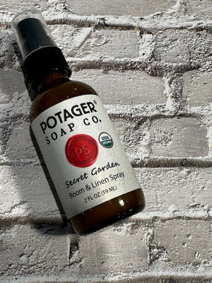 Potager Soap Co. Secret Garden organic room and linen spray in a 2 oz amber glass bottle on a white brick background.