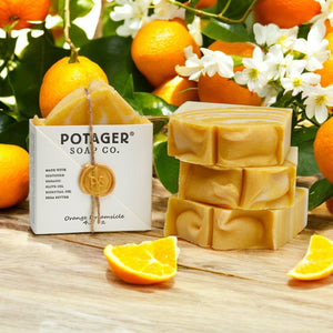 Potager Soap Co. Orange Dreamsicle soap bars with packaging on a wooden surface with oranges and ylang ylang flowers